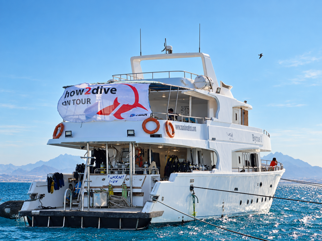 White dive boat with "how2dive ON TOUR" banner on blue sea, with a mountain coastline behind.