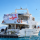 White dive boat with "how2dive ON TOUR" banner on blue sea, with a mountain coastline behind.