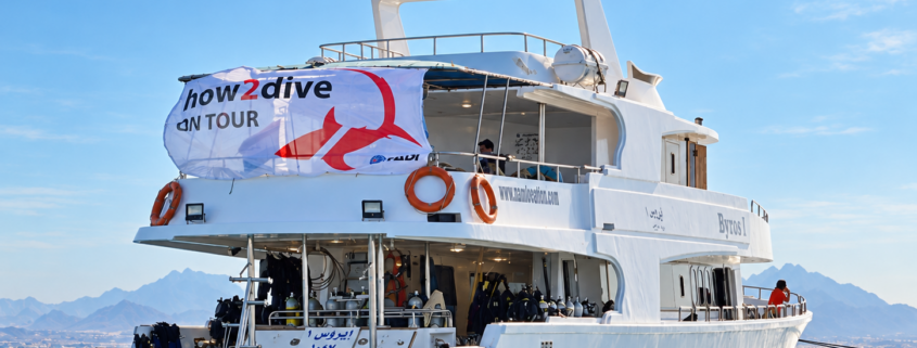 White dive boat with "how2dive ON TOUR" banner on blue sea, with a mountain coastline behind.